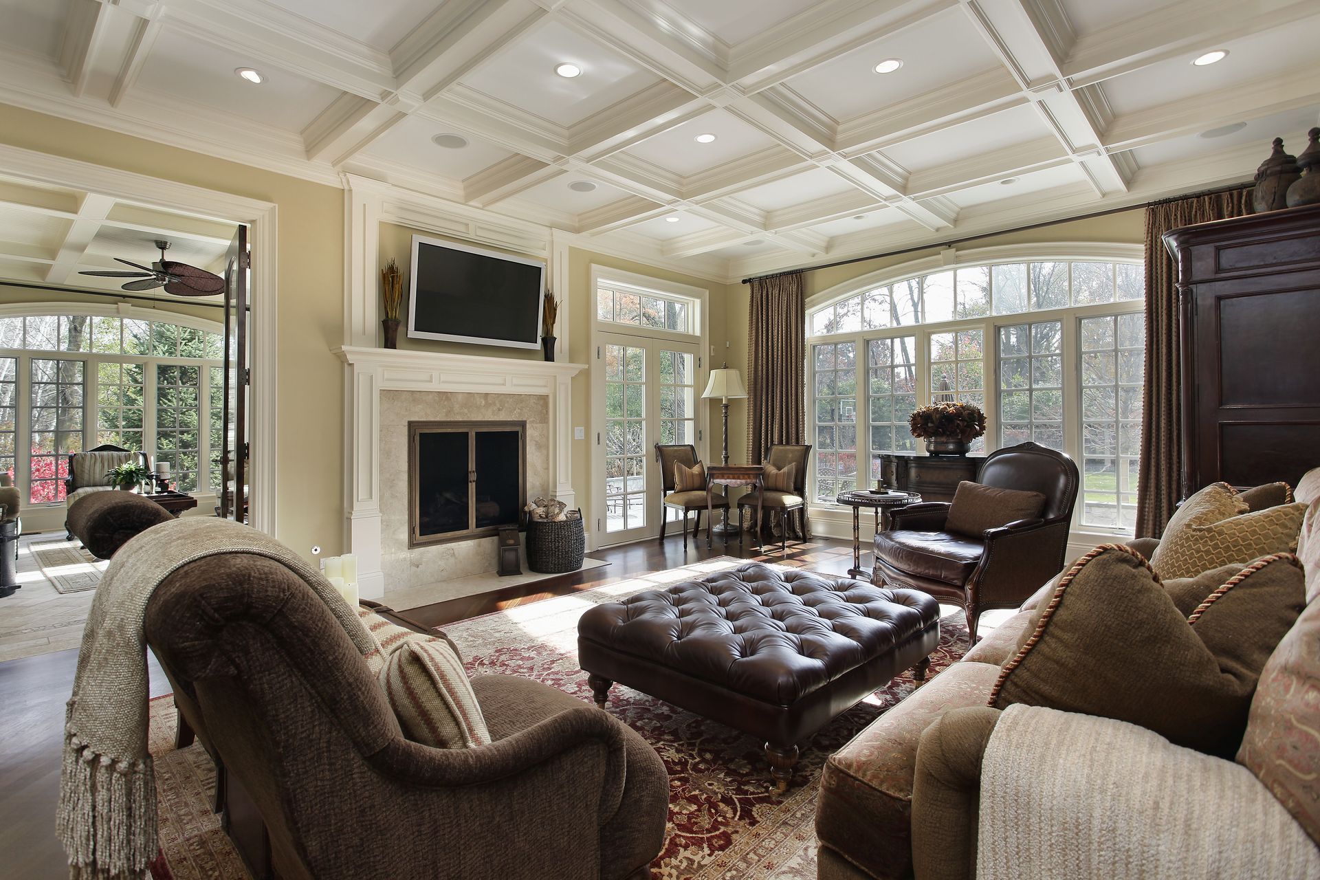 A luxury living room with a coffered ceiling, fireplace, leather ottoman, and large windows with garden views.