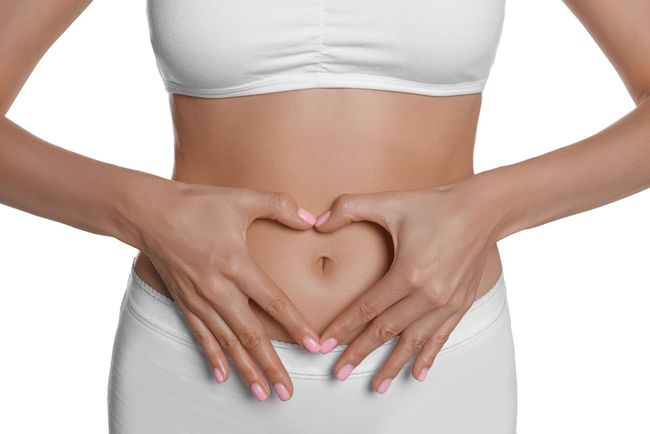Hände, die über einem nackten Bauch vor weißem Hintergrund ein Herz formen