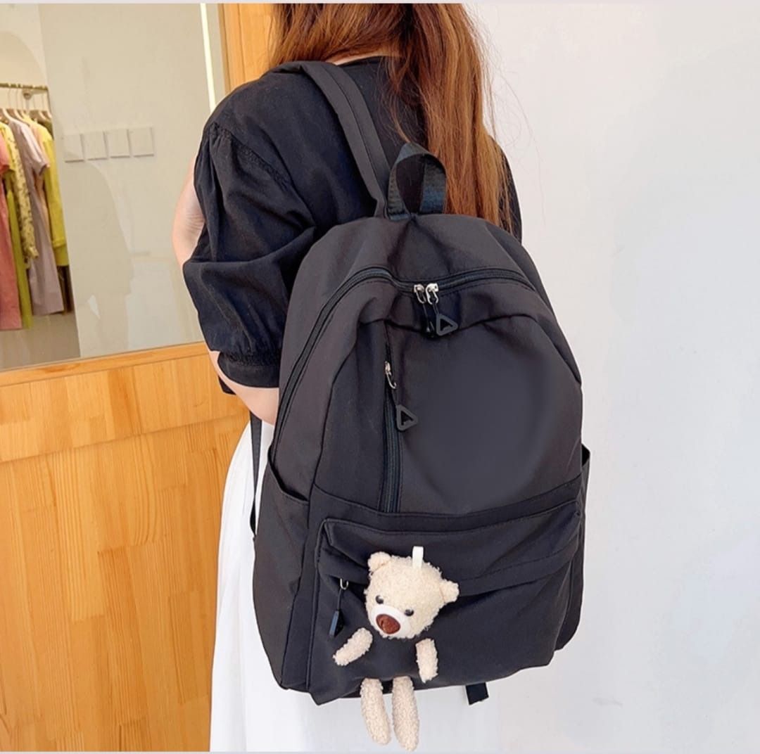 Una mujer lleva una mochila negra con un osito de peluche atado a ella.