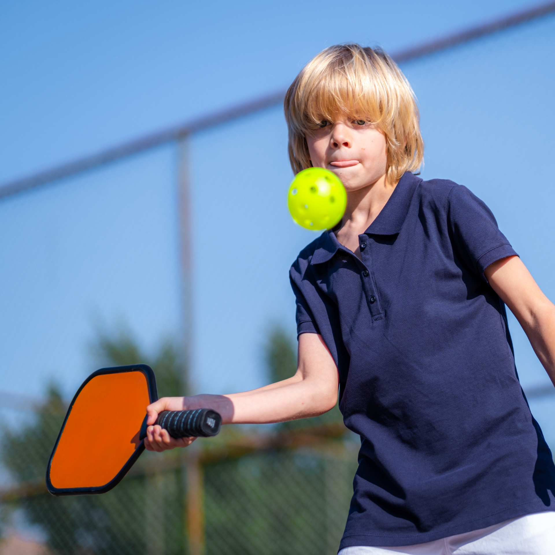 A young boy is holding a paddle and a tennis ball in his mouth
