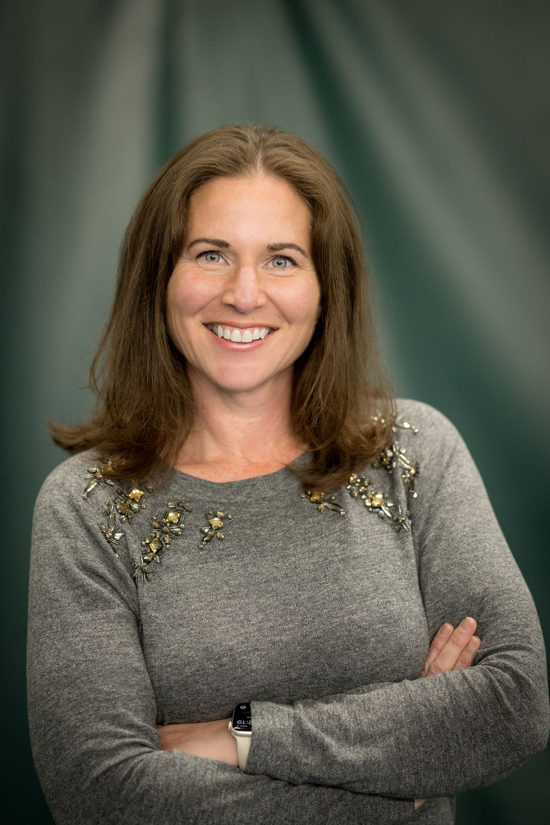A woman in a grey sweater is smiling with her arms crossed.