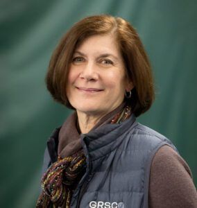A woman wearing a vest and scarf is smiling for the camera.