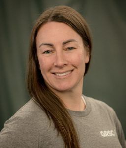 A woman with long hair is wearing a grey shirt and smiling.