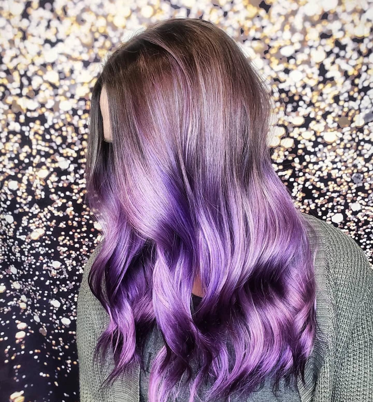 A person with long, wavy hair transitioning from a dark brown root to a vibrant, metallic purple ombre finish.