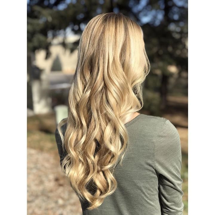 A person from the back with long, wavy blonde hair wearing a grey long-sleeved shirt outdoors in soft sunlight.