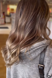 A person with shoulder-length, wavy hair featuring brown and blonde highlights, wearing a gray hooded sweatshirt.