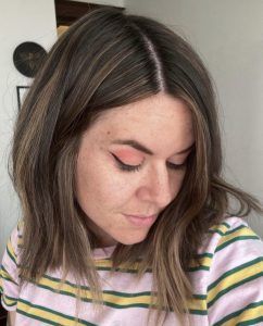 A person with shoulder-length, light brown wavy hair and a pink striped shirt, looking downward.