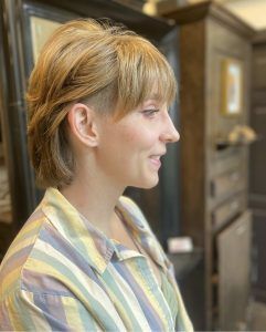 A side profile of a person with blonde hair in a layered cut with shaved temples, wearing a striped, collared shirt.