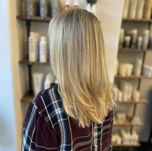 A person with long, blonde layered hair stands in a salon in front of shelves filled with hair care products.