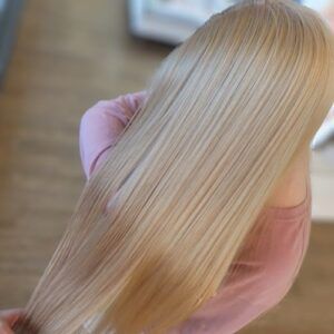 A close-up of straight, light-blonde hair flowing down the back of a person wearing a pale pink long-sleeve shirt.