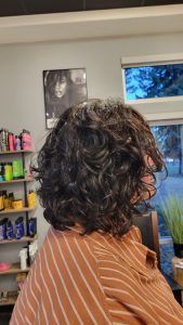 A person with dark, wavy-to-curly hair seen from behind inside a hair salon with product shelves in the background.