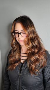 A person with long, wavy, brown highlighted hair wears black-rimmed glasses and a black jacket against a gray wall.