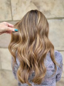 A person with long, wavy brown hair featuring blonde highlights, seen from behind against a textured stone wall.