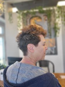 A side view of a person with a short, textured brown haircut, sitting in a hair salon with greenery in the background.