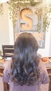 A person with long, dark, wavy hair stands in a salon with a branded 