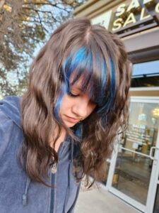 A person with long, wavy brown hair and blue highlights in their bangs standing in front of a salon.