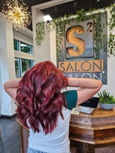 A person with vibrant, wavy red hair stands at a salon reception desk beneath an S2 Salon sign.