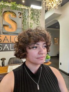 A person with shoulder-length curly brown hair and bangs stands in a hair salon with a neutral expression.