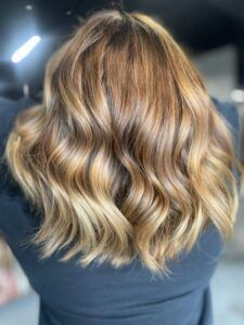 Medium-length hair styled in soft, loose waves with golden-brown highlights and darker roots.