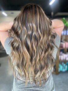 A person with long, wavy brown hair featuring blonde highlights, styled in loose curls, viewed from behind.