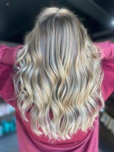 A back view of long, wavy hair with blonde balayage highlights, styled in loose curls against a maroon background.