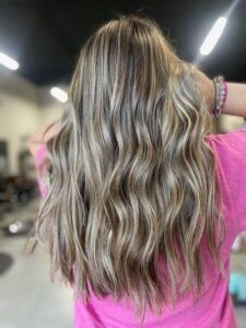 A person with long, wavy blonde highlighted hair viewed from behind, wearing a pink shirt in a salon setting.