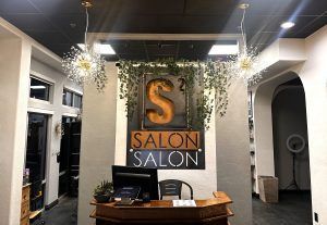 Reception desk at S2 Salon featuring a prominent gold logo, hanging greenery, and two decorative starburst pendant lights.
