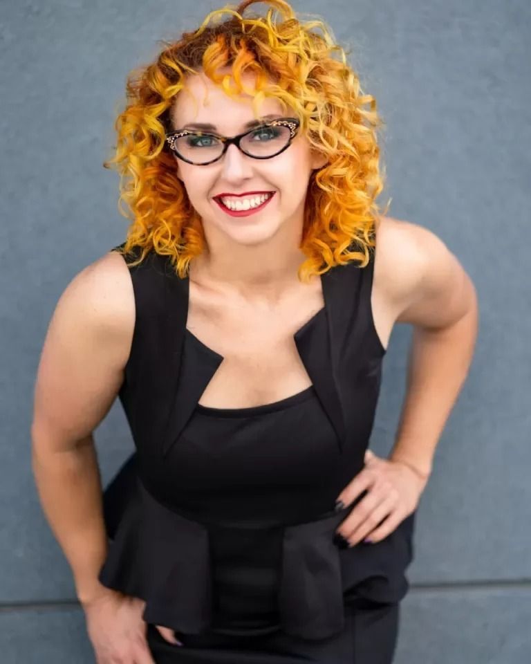 A smiling woman with vibrant curly hair and glasses wears a black dress against a plain grey background.