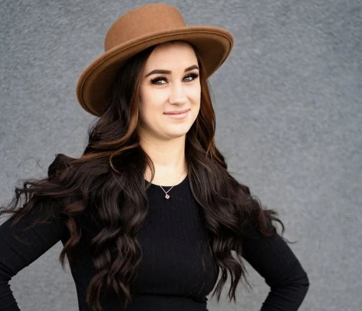 A person with long, dark wavy hair wearing a brown hat and black long-sleeve shirt poses against a grey background.
