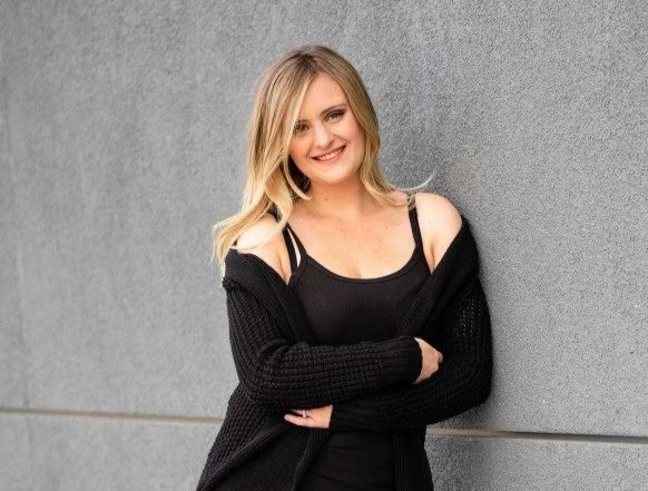 A smiling person with long blonde hair, wearing a black tank top and cardigan, leaning against a gray textured wall.