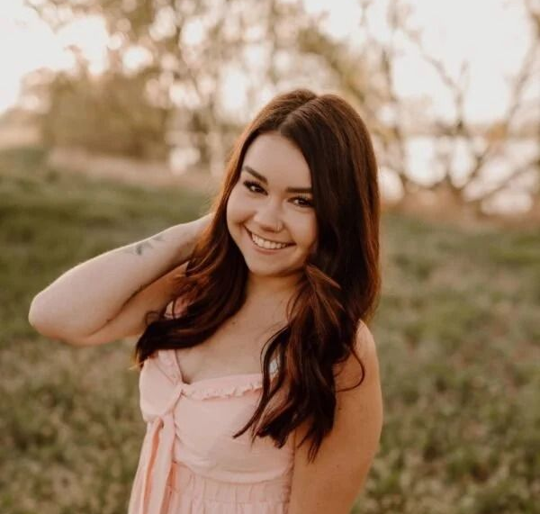 A person with long brown hair, wearing a light pink top, smiles while posing outdoors in a grassy field at sunset.