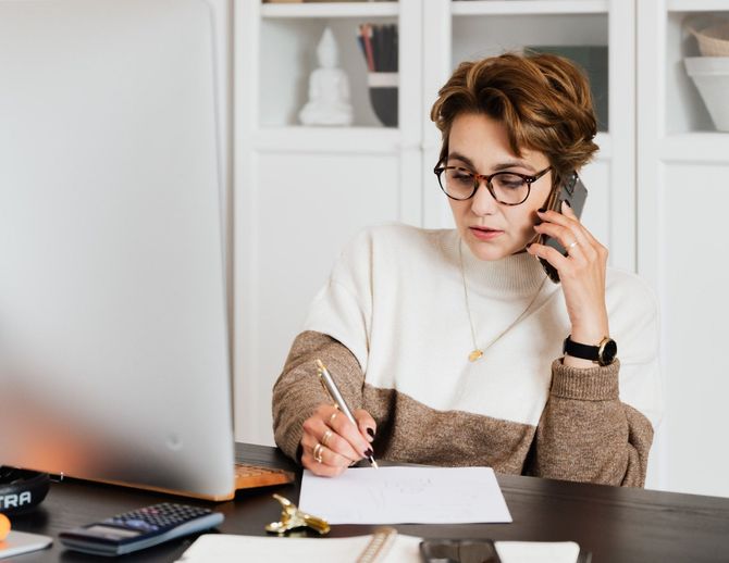 A woman is sitting at a desk talking on a cell phone and writing on a piece of paper.
