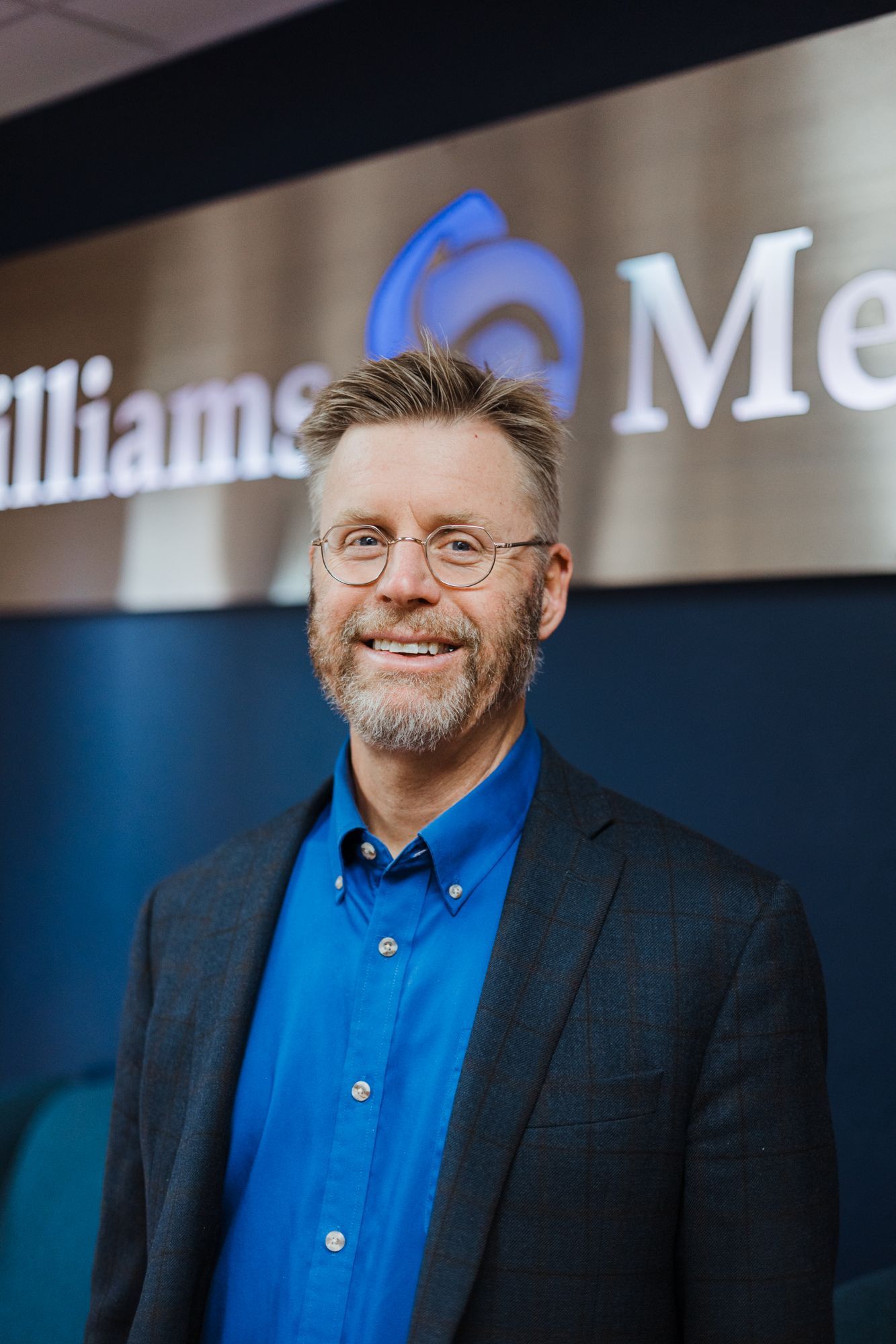 A man in a suit and blue shirt is standing in front of a sign.