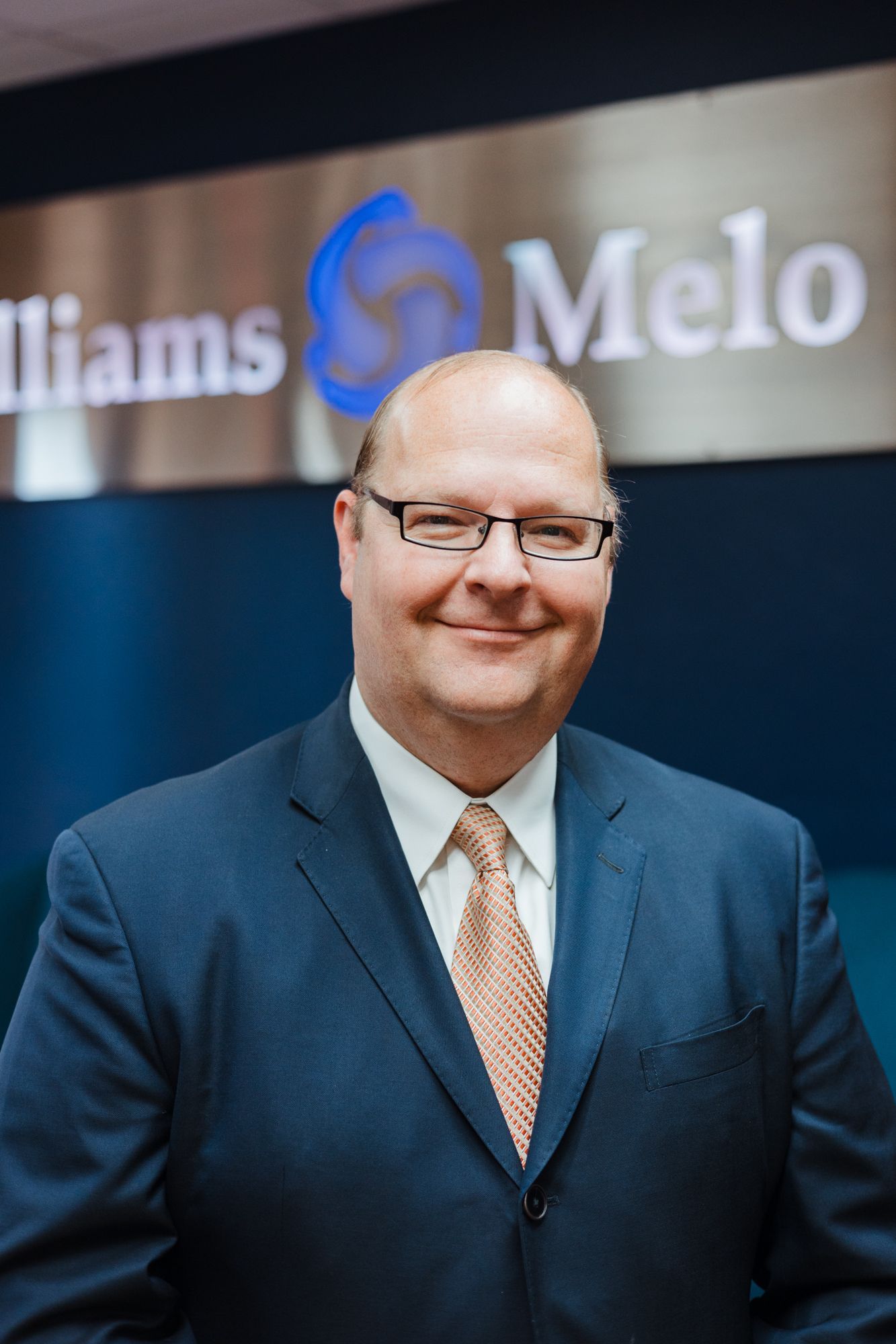 A man in a suit and tie is smiling in front of a williams melo sign.