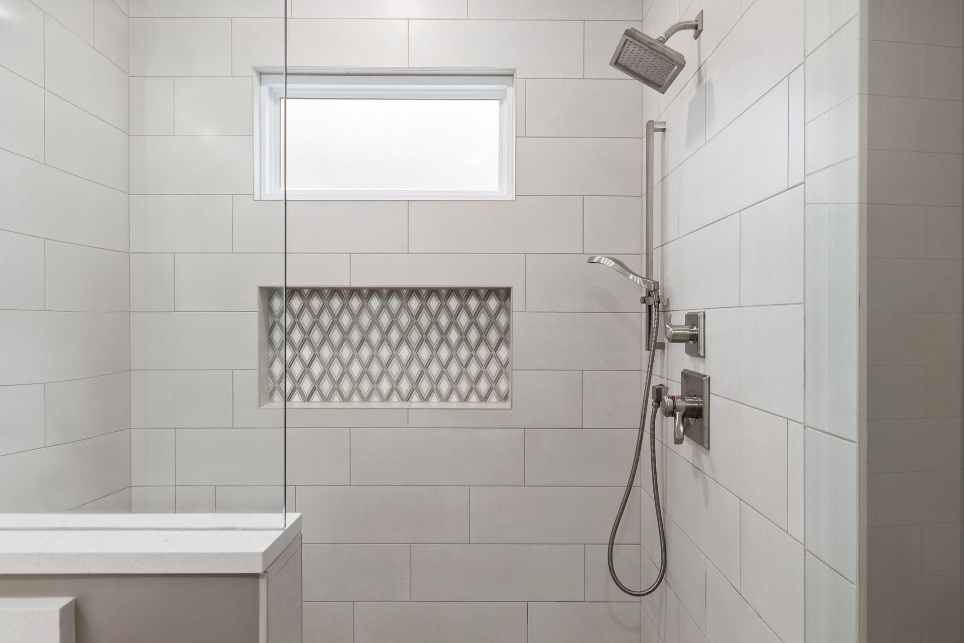 A bathroom with white tiles , a shower , a window and a tub.