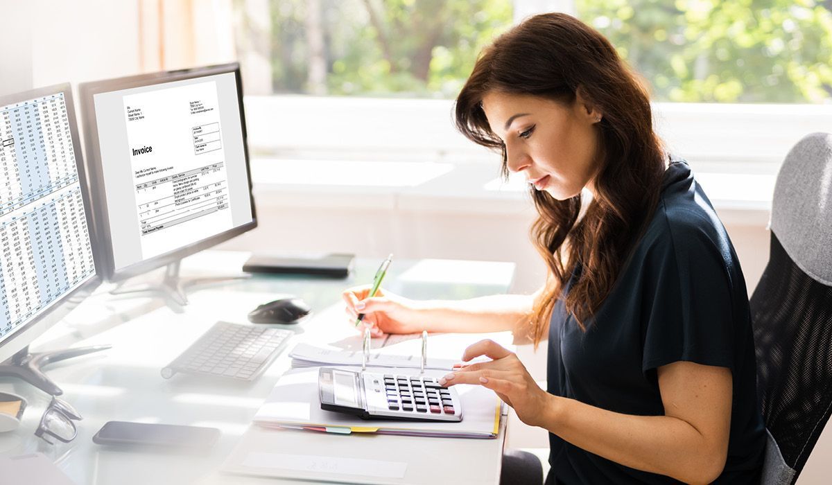 Woman at a bright desk, writing in a notebook beside a laptop and calculator.