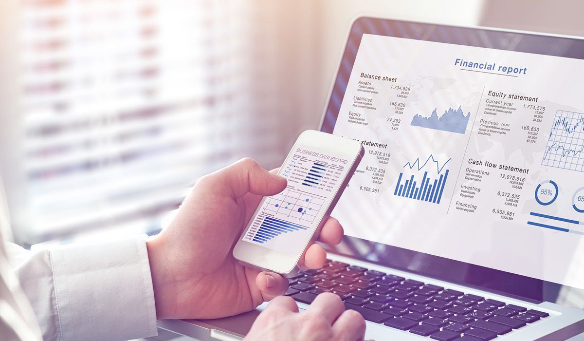 Hand holding smartphone beside laptop showing analytics dashboards and charts