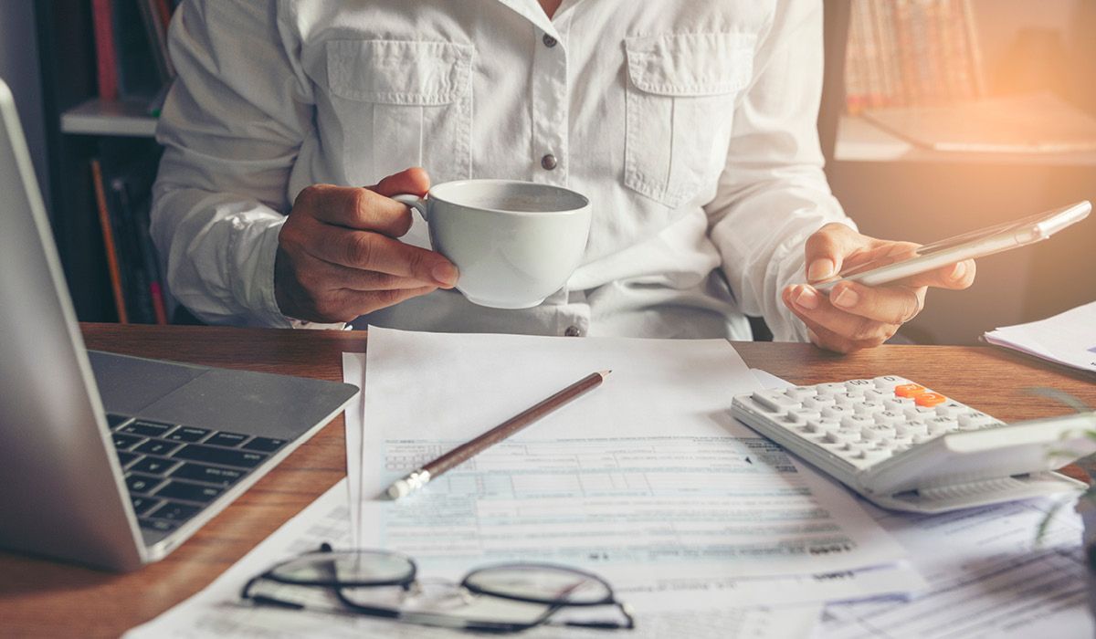 Person at a desk with laptop, coffee cup, calculator, and paperwork in a bright office setting