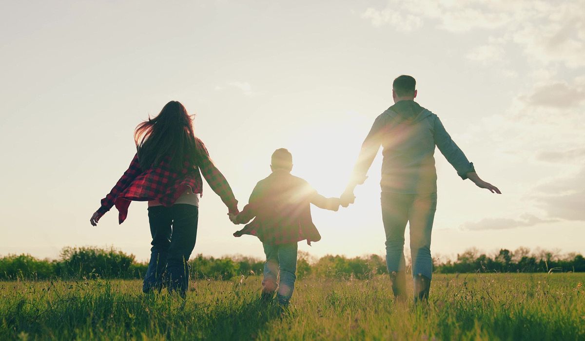 Three people holding hands and walking through a sunlit field at sunset.