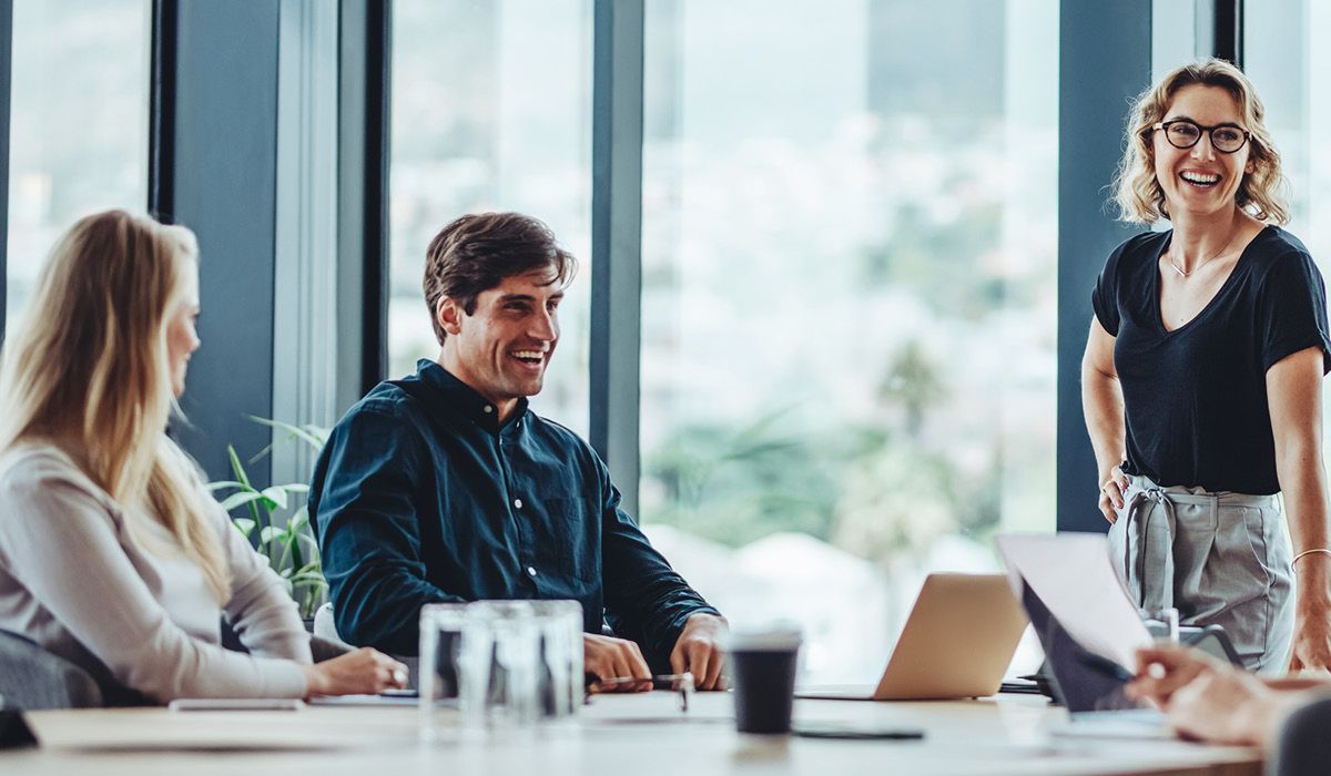 Three coworkers in a bright office meeting, talking around a conference table with a laptop.