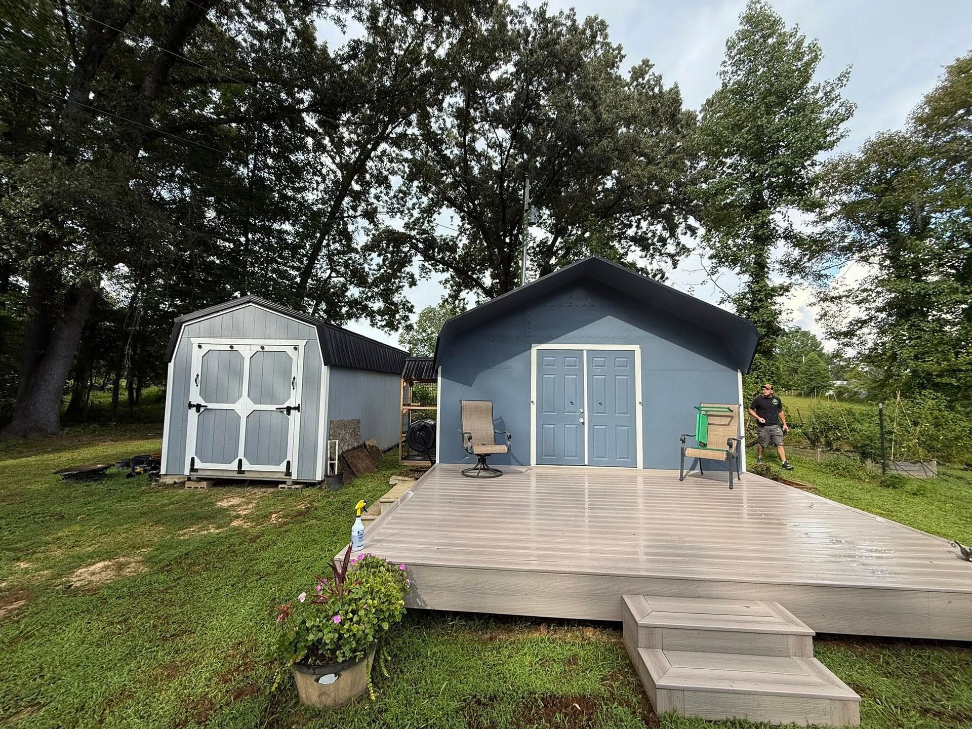 Two sheds in a grassy, wooded yard; one is gray with a large deck and two chairs, the other is white with double doors.