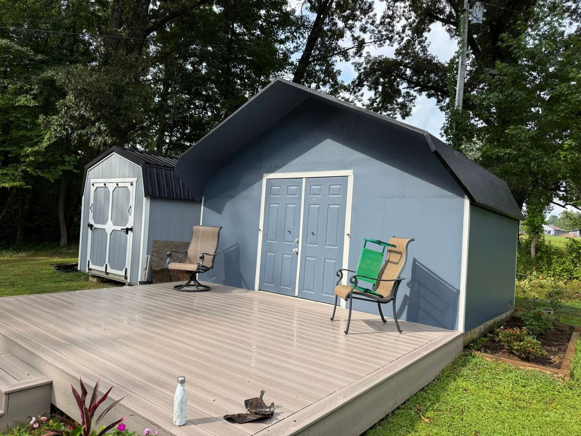 A grey storage shed with double doors and a smaller shed behind it, both situated on a large, light-colored patio deck.