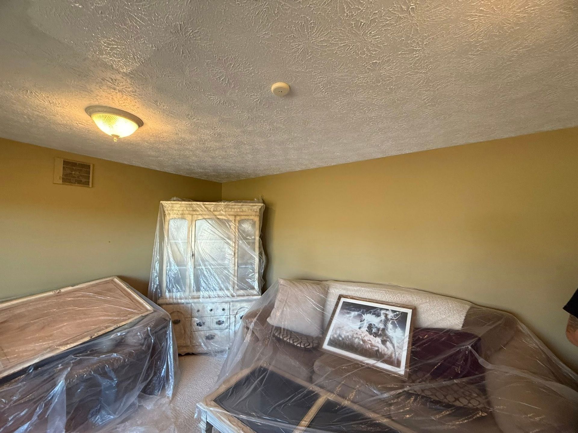 A room with beige walls, a popcorn ceiling, a light fixture, a covered hutch, and furniture covered in plastic sheeting.