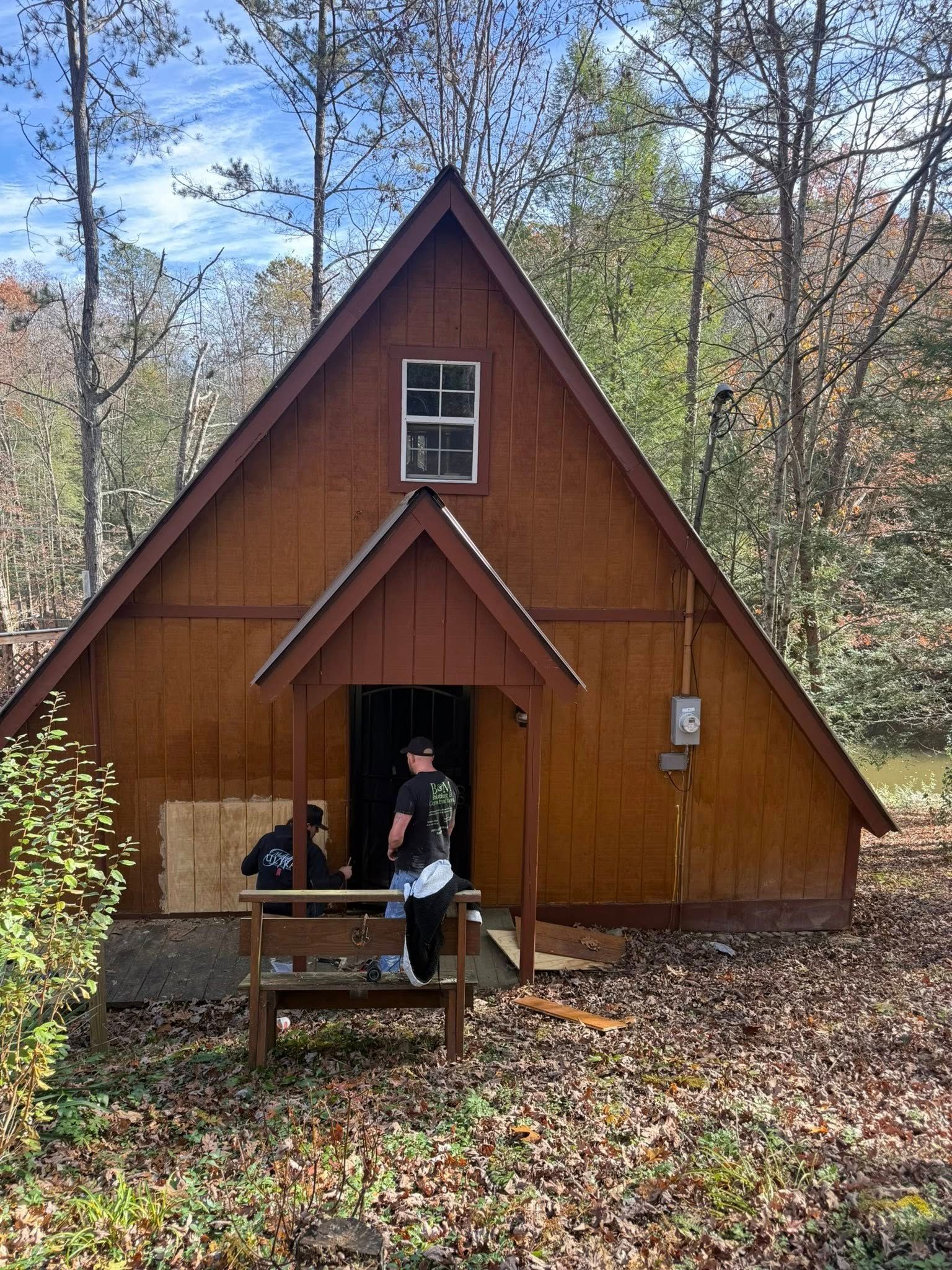 Two people stand at the entrance of a rustic, triangular A-frame cabin in a wooded, leaf-covered area.