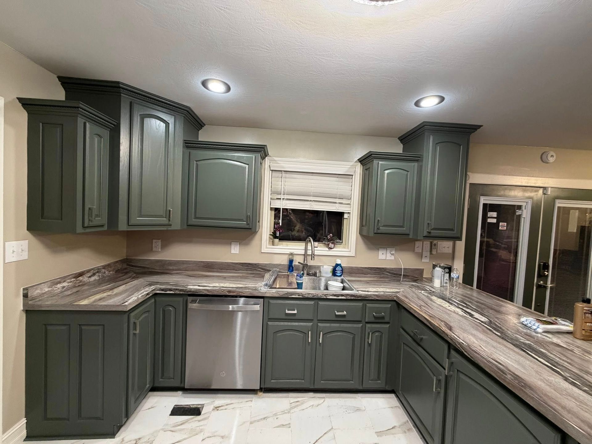 A U-shaped kitchen with dark sage cabinets, marble-patterned countertops, a stainless steel dishwasher, and tile flooring.