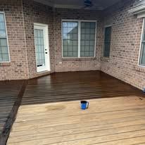 A wooden deck against a brick house shows a clear contrast between a dark, stained section and an unfinished light area.