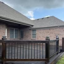 A brown wooden deck with a railing attached to the back of a brick house under a blue sky with scattered clouds.