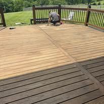 A person staining a wooden deck, showing a distinct contrast between the light, unfinished wood and the dark-stained area.