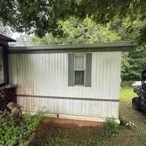 Exterior wall of a mobile home with vertical white siding, gray trim, and one window with dark shutters.
