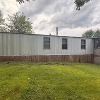 A light-gray mobile home exterior with a brown wood-paneled foundation and three windows, situated on a grassy lot.